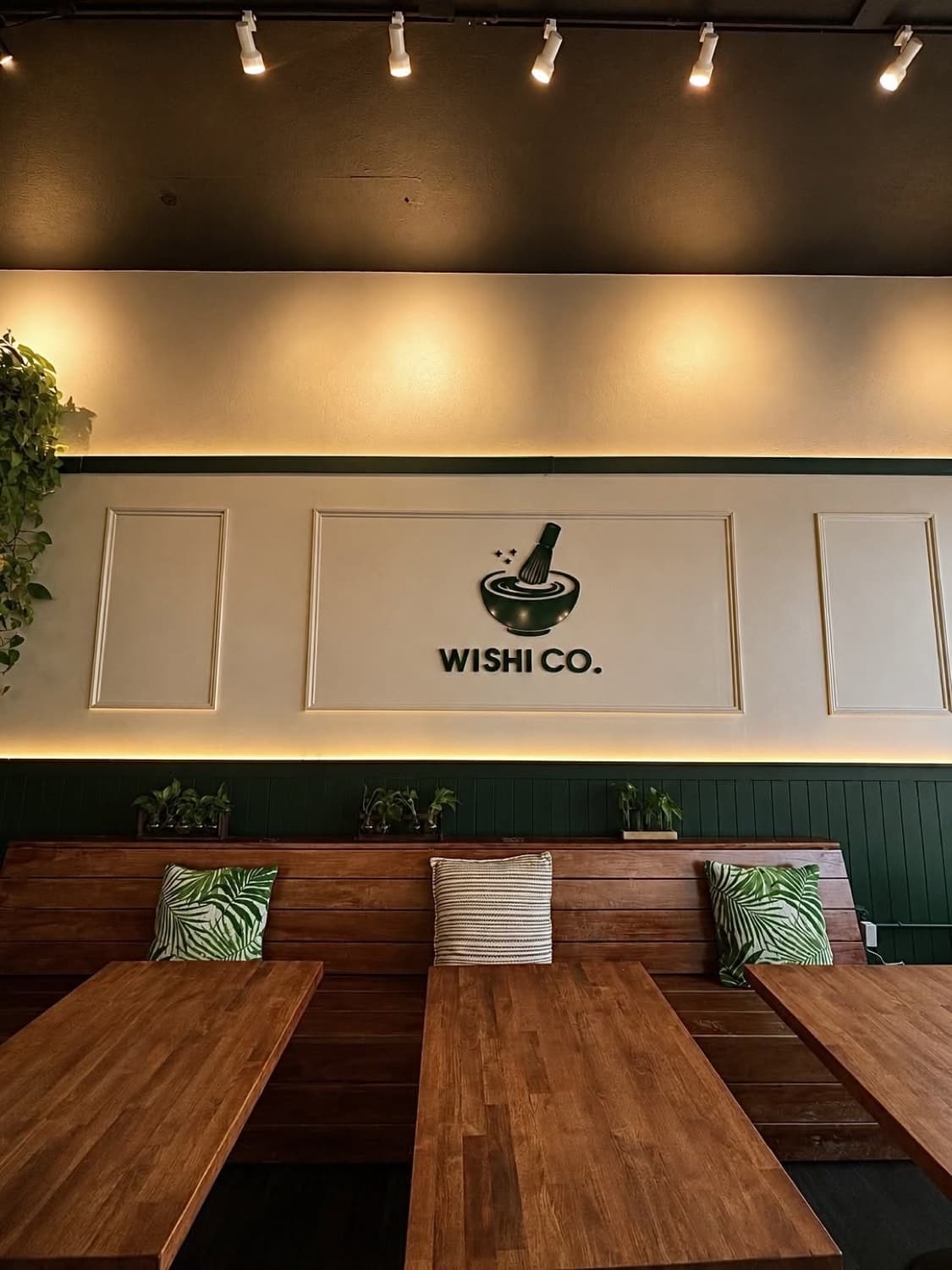 Inside Wishi Co. — communal tables and plants beneath the matcha-whisk mark.
